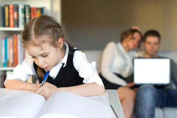 Little girl doing homework, while her parents resting on the sofa. Stock Photo