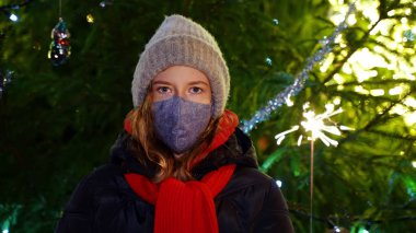 Elinde maytapla maske takan üzgün küçük kız. Covid Noelleri.