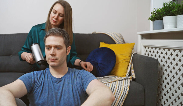 Женщина массирует мужчину с помощью массажного перкуссионного устройства дома.