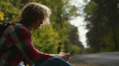 Yolun kenarında cep telefonuyla oturan erkek turist.