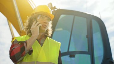 Sert şapkalı kazıcı operatörü akıllı telefonla konuşuyor..
