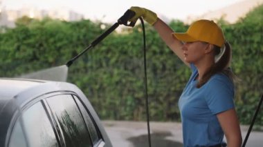 Üniformalı bir kadın yüksek basınçlı su kullanarak araba temizliyor. Araba yıkama servisi.