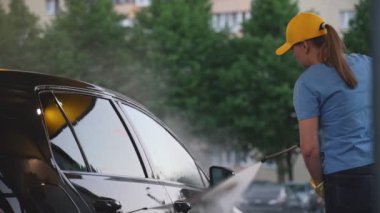 Üniformalı bir kadın yüksek basınçlı su kullanarak araba temizliyor. Araba yıkama servisi.