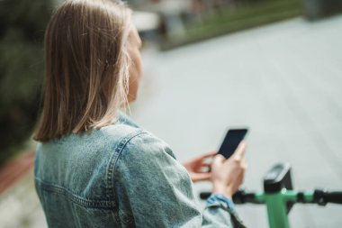 Sokakta elektrikli scooterın kilidini açmak için akıllı telefondaki uygulamayı kullanan kadın..