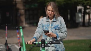 Sokakta elektrikli scooterın kilidini açmak için akıllı telefondaki uygulamayı kullanan kadın..