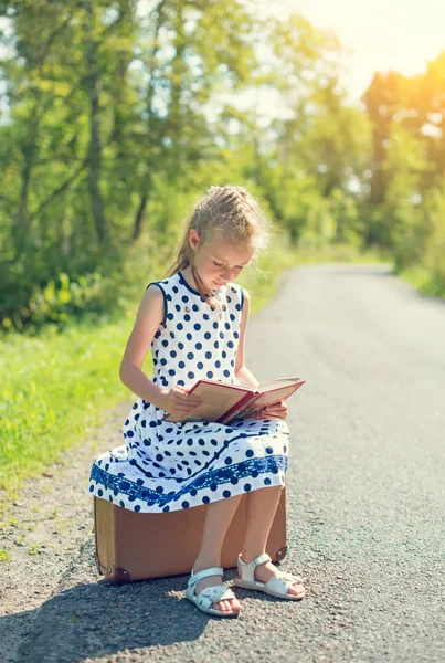 Niña sentada en una maleta y leyendo un libro. Esperando el transporte . —  Fotos de Stock