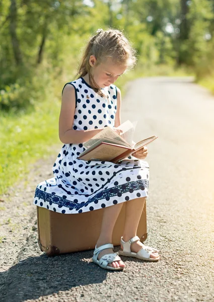 Ragazzina seduta sulla valigia e che legge un libro. In attesa di trasporto . — Foto Stock