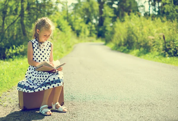 Petite fille assise sur une valise et lisant un livre. En attente de transport . — Photo