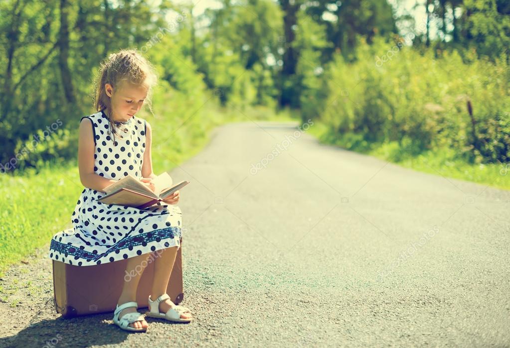 Little girl sitting on suitcase and reading book. Waiting for transport