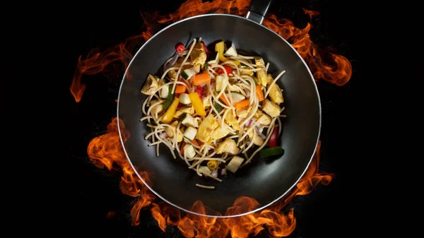 Asian noodles in wok pan, flames on background. High angle view of meat preparation, studio shot.