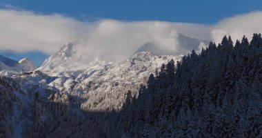 Avusturya 'daki karlı dağların sabah sisinin içinden yükselen bir buzulun havadan çekilmiş fotoğrafı. Mavi gökyüzü, güneş ışığı ve ön plandaki kozalaklı ormanlar huzurlu bir kış ortamı yaratır..