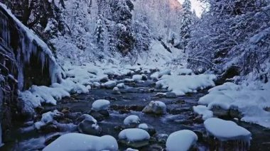 Karla kaplı bir akarsu üzerinde hafif kar yağışı olan huzurlu bir yavaş çekim uçuşu. Akan buzlu su, donmuş kayalar ve kar yağışı dinlenmek ve doğa sahneleri için sakin bir kış atmosferi yaratır..