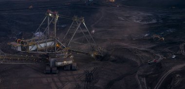 Toz ve dramatik ışıkla çevrili açık bir kömür madeninde çalışan devasa maden makinelerinin aşırı yüksek çözünürlüklü panoramik görüntüsü. Büyük ölçekli çıkarma, enerji üretimi ve insan etkisinin görüldüğü güçlü sanayi sahnesi.