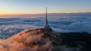 Jested Tower ve Hotel 'in hava manzarası gün doğumunda bulutların üzerinde yükseliyor. Sıcak altın ışık ve sabah sisi Çek Cumhuriyeti 'ndeki Jizera Dağları üzerinde gerçeküstü, barışçıl bir atmosfer yaratır..