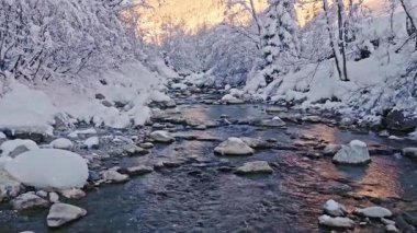 Karla kaplı bir akarsu üzerinde hafif kar yağışı olan huzurlu bir yavaş çekim uçuşu. Akan buzlu su, donmuş kayalar ve kar yağışı dinlenmek ve doğa sahneleri için sakin bir kış atmosferi yaratır.
