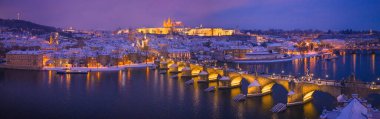 Prag 'ın yüksek çözünürlüklü kış manzarasında Charles Bridge, Prag Kalesi ve Vltava Nehri alacakaranlıkta. Sıcak ışıklarla aydınlatılmış tarihi şehri kaplayan kar büyülü bir Avrupa kış atmosferi yaratır..