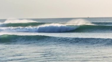 Ağır çekimde güçlü bir okyanus dalgası etrafında sinemalar uçuşuyor, arka planda dalgalanan dalgalar ve sörfçülerle bir sörfçü cenneti yakalıyor. Doğal deniz atmosferi, enerji ve sörf kültürü özgürlüğü.