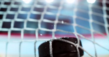 Buz hokeyi diski, çarpışma anında patlayan buz parçacıkları ve arka planda bulanık bir kış stadyumu ve buz pateni pisti ile 1000 fps hızla gol ağına çarpıyor..