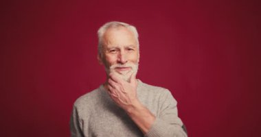 Gülümseyen son sınıf öğrencisinin gri sakala dokunup kameraya baktığı portresi, kırmızı arka planda yalnız poz veren modaya uygun yaşlı erkek model. Reklam kavramı