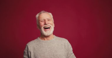 Gülümseyen, mutlu ve yaşlı bir adamın portresi. Kameraya bakıp gülüyor, kırmızı arka planda izole bir şekilde poz veriyor. Arkadaş canlısı emekli erkek pozu. Pozitif duygu kavramı