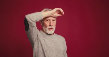 Kırmızı arka plandaki kameraya bakarak alnını silen yorgun, üzgün yaşlı adamın portresi. Stresli, olgun bir erkek. Olumsuz duygular konsepti