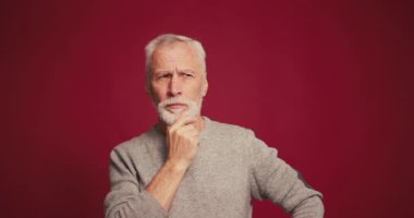 Gülümseyen, mutlu yaşlı sakallı adamın iyi bir fikri var, kırmızı arka planda izole edilmiş kameraya işaret ediyor. Beyin fırtınası konsepti