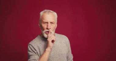 Ciddi, gri saçlı, dudak kenarında parmak tutan yaşlı bir adamın portresi kırmızı arka planda izole edilmiş. Sessizlik kavramı