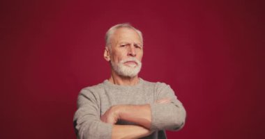 Ciddi, sakallı, çapraz kollu yaşlı bir adam kırmızı arka planda izole edilmemiş bir kafayla kameraya bakıyor. Duygular konsepti