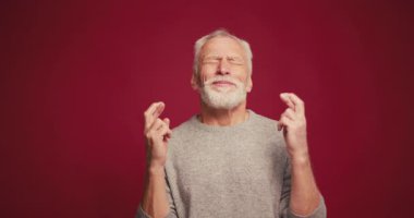 Mutlu, gülümseyen gri saçlı, parmakları çapraz olan olgun bir adam kırmızı arka planda izole edilmiş bir şekilde dilek diliyor. Umut kavramı