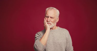 Üzücü, üzgün yaşlı adamın elinde çek var. Kırmızı arka planda duran kameraya bakarken diş ağrısı çekiyor. Diş sağlığı konsepti