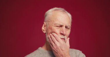 Üzücü, üzgün yaşlı adamın elinde çek var. Kırmızı arka planda duran kameraya bakarken diş ağrısı çekiyor. Diş sağlığı konsepti