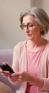 Olgun 60 'lı yaşlarda kadın, akıllı telefon kullanan orta yaşlı bayan müşteri cep telefonu uygulaması, mesaj gönderme, cep telefonu teknolojisi cihazında arama önerileri evdeki koltukta oturuyor..