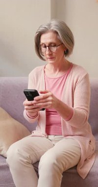 Olgun 60 'lı yaşlarda kadın, akıllı telefon kullanan orta yaşlı bayan müşteri cep telefonu uygulaması, mesaj gönderme, cep telefonu teknolojisi cihazında arama önerileri evdeki koltukta oturuyor..