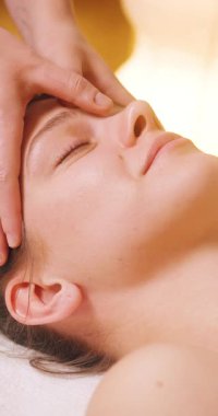 Spa Salonu 'nda yatakta uzanan ve yüz masajından zevk alan mutlu genç kadın. Lenfatik drenaj ve kaldırma efekti için kadın müşteri yüzüne masaj yapan kozmetik uzmanı. Güzellik, Cilt Bakımı.