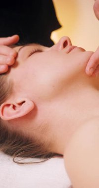 Spa Salonu 'nda yatakta uzanan ve yüz masajından zevk alan mutlu genç kadın. Lenfatik drenaj ve kaldırma efekti için kadın müşteri yüzüne masaj yapan kozmetik uzmanı. Güzellik, Cilt Bakımı.