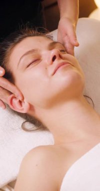 Spa Salonu 'nda yatakta uzanan ve yüz masajından zevk alan mutlu genç kadın. Lenfatik drenaj ve kaldırma efekti için kadın müşteri yüzüne masaj yapan kozmetik uzmanı. Güzellik, Cilt Bakımı.
