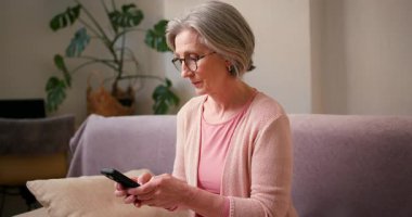 Olgun 60 'lı yaşlarda kadın, akıllı telefon kullanan orta yaşlı bayan müşteri cep telefonu uygulaması, mesaj gönderme, cep telefonu teknolojisi cihazında arama önerileri evdeki koltukta oturuyor..