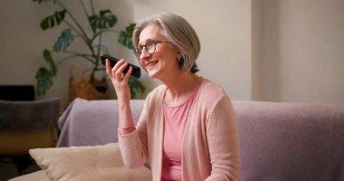 Olgun 60 'lı yaşlarda kadın, orta yaşlı bayan müşteri akıllı telefon tutuyor cep telefonu uygulaması kullanıyor sesli mesaj dinliyor ve evdeki koltukta oturan cep telefonu teknolojisi cihazından ses kaydediyor..