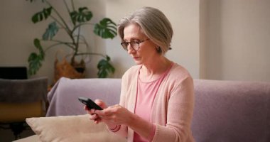 Olgun 60 'lı yaşlarda kadın, akıllı telefon kullanan orta yaşlı bayan müşteri cep telefonu uygulaması, mesaj gönderme, cep telefonu teknolojisi cihazında arama önerileri evdeki koltukta oturuyor..