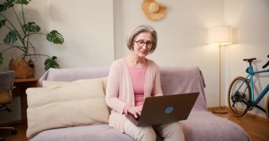 Evde çalışan 60 'lı yaşlardaki olgun bayan öğrenci evdeki kanepede oturan dizüstü bilgisayar teknolojisi cihazıyla çalışıyor..