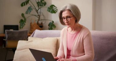 Evde çalışan 60 'lı yaşlardaki olgun bayan öğrenci evdeki kanepede oturan dizüstü bilgisayar teknolojisi cihazıyla çalışıyor..