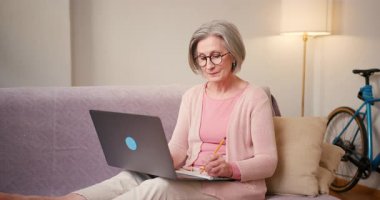 Evde çalışan 60 'lı yaşlardaki olgun bayan öğrenci evdeki kanepede oturan dizüstü bilgisayar teknolojisi cihazıyla çalışıyor..