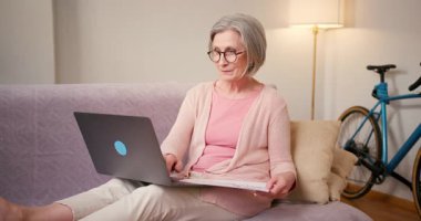 Evde çalışan 60 'lı yaşlardaki olgun bayan öğrenci evdeki kanepede oturan dizüstü bilgisayar teknolojisi cihazıyla çalışıyor..