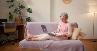 Evde çalışan 60 'lı yaşlardaki olgun bayan öğrenci evdeki kanepede oturan dizüstü bilgisayar teknolojisi cihazıyla çalışıyor..