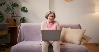 Yaşlı, olgun 60 'lı yaşlarda bir kadın, orta yaşlı bir kadın. Video görüşmesi yapıyor. Evde koltukta otururken dizüstü bilgisayarla gülümsüyor..