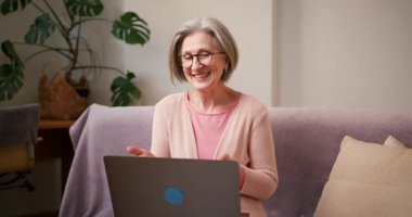 Yaşlı, olgun 60 'lı yaşlarda bir kadın, orta yaşlı bir kadın. Video görüşmesi yapıyor. Evde koltukta otururken dizüstü bilgisayarla gülümsüyor..