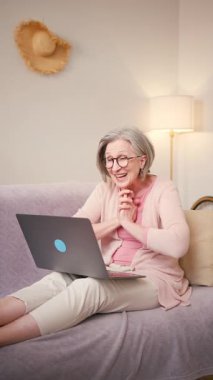 Yaşlı, olgun 60 'lı yaşlarda bir kadın, orta yaşlı bir bayan müşteri evde koltukta otururken dizüstü bilgisayarla gülerek video görüşmesi yapıyor. Dikey video