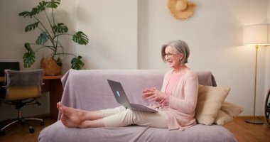 Yaşlı, olgun 60 'lı yaşlarda bir kadın, orta yaşlı bir kadın. Video görüşmesi yapıyor. Evde koltukta otururken dizüstü bilgisayarla gülümsüyor..
