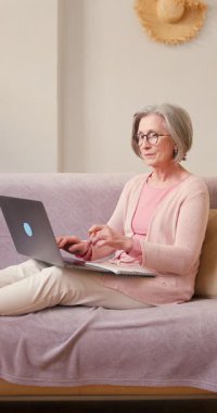 Evde çalışan 60 'lı yaşlardaki olgun bayan öğrenci evdeki kanepede oturan dizüstü bilgisayar teknolojisi cihazıyla çalışıyor..