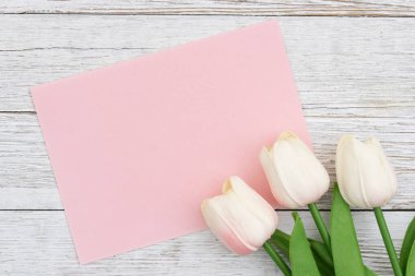 Beyaz laleler ve kırsal ahşap üzerinde pembe kart selamlama, davet ya da mevsimsel mesaj tasarımı için yumuşak bahar kompozisyonu oluşturur.